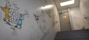 This image shows a stairwell with various hand-drawn artworks on the walls. One prominent illustration features two characters resembling Pikmin (one orange and one blue) carrying a large fruit. The rest of the wall has several black-and-white sketches, including a detailed drawing of a person with a sword and another figure in a suit. The setting appears to be an industrial or institutional stairwell at College of the Canyons third floor stariwell in the Menrty Hall Building.